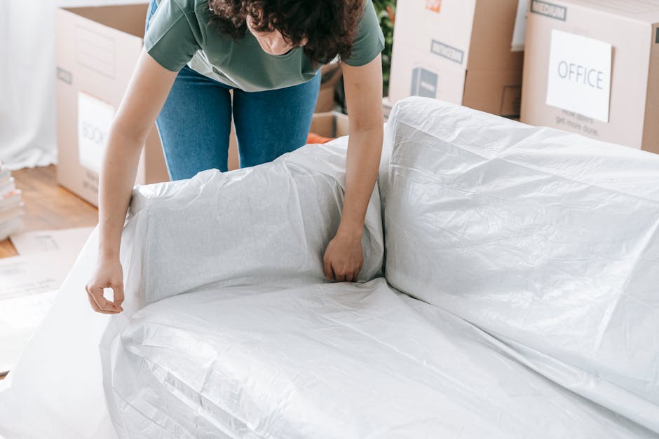 A person with curly hair wearing a green T-shirt and blue jeans is seen preparing a large upholstered sofa for a home relocation inside a room. They are carefully wrapping the sofa with protective white plastic sheeting, ensuring the fabric and cushions are covered for transport. The sofa is positioned near the doorway, with cardboard boxes and packing materials visible around the room, indicating an ongoing packing and moving process. The scene includes a wooden floor and natural lighting, with stairs visible in the background. This image illustrates the packing stage of a furniture transport service provided by Removal Van Marylebone, focusing on the careful handling of bulky furniture during a house removal in Marylebone, aligning with professional relocation practices.