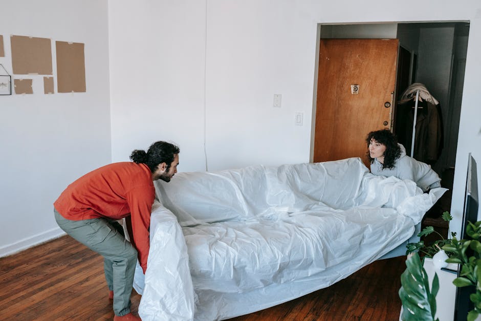 A person with curly hair wearing a green T-shirt and blue jeans is seen preparing a large upholstered sofa for a home relocation inside a room. They are carefully wrapping the sofa with protective white plastic sheeting, ensuring the fabric and cushions are covered for transport. The sofa is positioned near the doorway, with cardboard boxes and packing materials visible around the room, indicating an ongoing packing and moving process. The scene includes a wooden floor and natural lighting, with stairs visible in the background. This image illustrates the packing stage of a furniture transport service provided by Removal Van Marylebone, focusing on the careful handling of bulky furniture during a house removal in Marylebone, aligning with professional relocation practices.