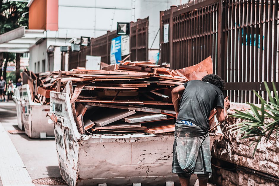 A man with curly dark hair wearing a black t-shirt and grey plaid shorts is standing outside a property, next to a large, white, worn-out skip filled with a variety of construction debris, including wooden planks, cardboard boxes, and plastic packaging materials. He appears to be inspecting or managing the contents of the skip, which is situated on a paved pavement adjacent to a brown metal fence and the building's exterior wall. Behind the skip, there are other vehicles and waste containers, indicating a waste disposal or renovation phase associated with home relocation or property refurbishment. The scene is well-lit with natural daylight, and in the background, there are additional pedestrians and urban elements. The presence of packaging materials and debris suggests ongoing packing, sorting, and loading processes typical of professional removals and furniture transport services carried out by companies like Removal Van Marylebone, supporting a smooth moving or relocation operation.