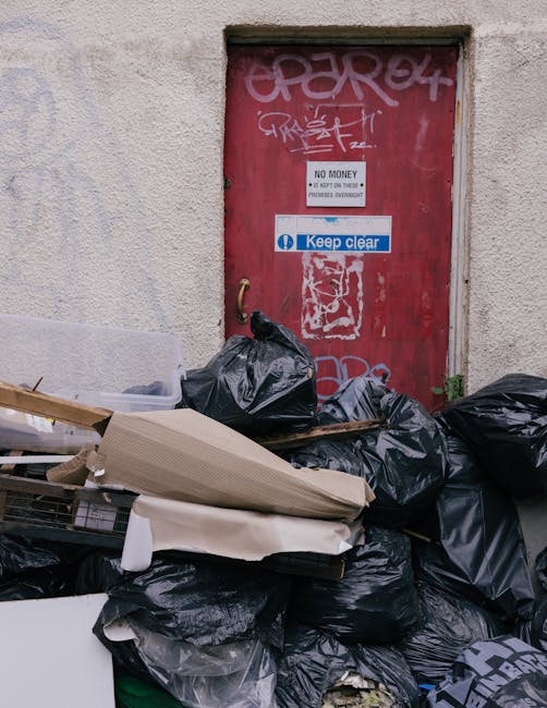 In front of a weathered, red metal door with graffiti and warning signs, a collection of black rubbish bags, cardboard boxes, and packing materials are piled on the pavement. The scene shows an outdoor area adjacent to a building, possibly part of a home or commercial property, used during packing or disposal in the house removal process. Behind the rubbish, there is a small concrete ledge, and the door features signs such as 'No Money' and 'Keep Clear.' The image captures the aftermath of packing for a home relocation, with various discarded packaging and debris typical during the loading and moving preparations facilitated by Removal Van Marylebone. The lighting appears natural, suggesting daytime, and the overall scene reflects a moment in the moving process involving waste clearance or preparatory staging for furniture transport and house removal services.
