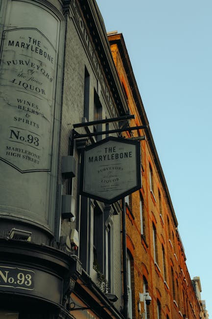 A photograph showing the exterior of a traditional brick building in Marylebone during daytime, with a black metal sign hanging from an ornate bracket displaying the name 'The Marylebone' and additional details about the location and businesses such as wine and spirits. The building features large windows, some with white trim, and the facade is partly illuminated by daylight. The sky above is clear with a light blue hue. This setting relates to house removals and home relocation services, as part of the typical environment where professional removals companies like Removal Van Marylebone operate, supporting logistics, packing, and furniture transport during house moves.