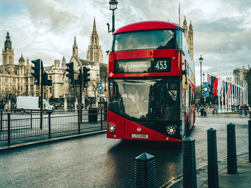 A red double-decker bus displaying the route number 453 and the destination Marylebone is approaching on a wet street near historic Gothic-style buildings with spires in the background. The bus is in motion, with its front on the paving, and there are traffic lights and a metal fence separating the roadway from a pedestrian walkway. To the right, a row of black bollards lines the sidewalk, and several pedestrians are visible in the distance, some walking and others standing near a flag display with Union Jack and other banners fluttering in the wind. The overcast sky reflects on the wet pavement, and a white van is seen moving further down the street. The scene captures a typical London urban environment, relevant to house relocations and furniture transport processes that Removal Van Marylebone provides for clients undertaking home removals or moving logistics in Marylebone.
