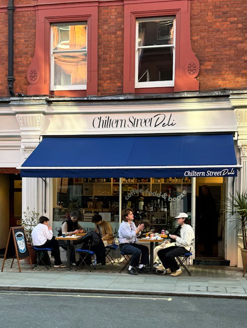 A daytime street view of Chiltern Street Deli located in a historic red-brick building with white decorative trim around the windows and a blue awning extending over the sidewalk. The shop's signage displays its name in elegant script, and the awning is branded with the same name on the side. Outside, five people are seated at small tables, enjoying food and drinks, with some engaging in conversation and others focused on their meal. The scene captures a casual, lively atmosphere typical of a popular local eatery, with sunlight illuminating the outdoor seating area. The image relates to the local environment that Removal Van Marylebone might be involved in when planning or executing house removals or furniture transport within the Marylebone area, taking into account busy street scenes with cafes and outdoor dining before or after a home relocation.