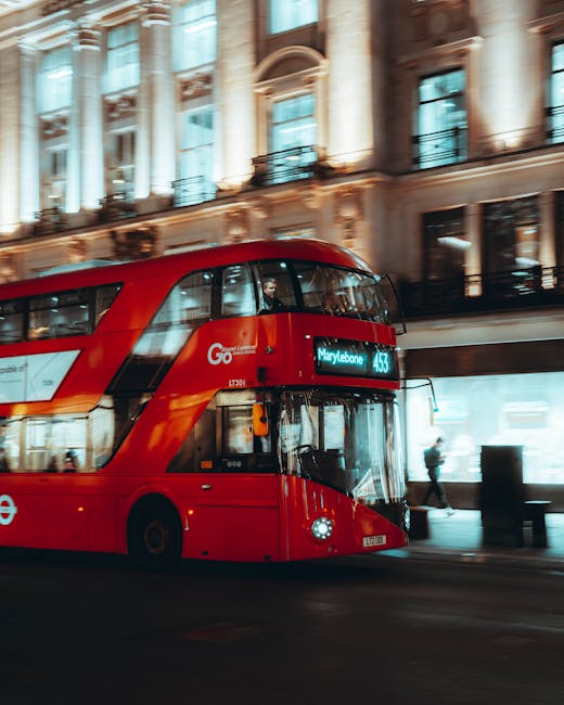 A red double-decker bus displaying the route number 453 and the destination Marylebone is approaching on a wet street near historic Gothic-style buildings with spires in the background. The bus is in motion, with its front on the paving, and there are traffic lights and a metal fence separating the roadway from a pedestrian walkway. To the right, a row of black bollards lines the sidewalk, and several pedestrians are visible in the distance, some walking and others standing near a flag display with Union Jack and other banners fluttering in the wind. The overcast sky reflects on the wet pavement, and a white van is seen moving further down the street. The scene captures a typical London urban environment, relevant to house relocations and furniture transport processes that Removal Van Marylebone provides for clients undertaking home removals or moving logistics in Marylebone.