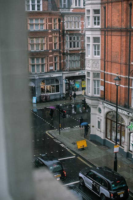View from a high window showing a rainy day scene in Marylebone, London, with pedestrians holding umbrellas crossing a wet street. The street is lined with red brick and white-painted buildings, some with bay windows. A dark grey van and a black car are parked along the curb, with a few people walking towards a shop entrance at the ground level of one building. A streetlamp, a yellow road sign, and a small parking area are visible on the pavement. Inside a residential property, glimpses of a partially open window frame suggest an ongoing home relocation or moving process managed by Removal Van Marylebone, near the entrance of a medical centre, illustrating the logistics involved in furniture transport or packing and moving activities in the area.