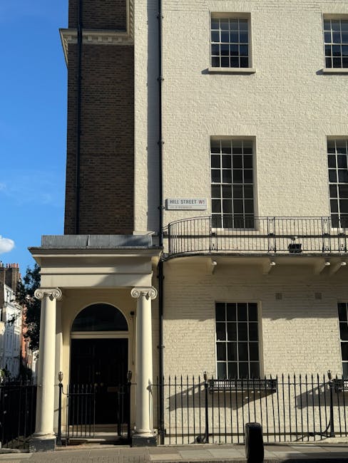 A street view of a three-story white building on Hill Street W1, featuring large rectangular windows with black frames and a small balcony with a black metal railing on the first floor. The building has a classical-style entrance with two white columns supporting a portico, surrounding a black front door. In front of the building, there is a black metal fence and a pedestrian pavement, with a small black wheelie bin visible near the curb. The sky above is clear and blue, indicating daytime. This setting is suitable for a house removal or relocation service, with space for a removal van to access the property for furniture transport and packing and moving activities, such as loading transport boxes and large household items. Removal Van Marylebone occasionally supports home relocation logistics on W1 streets, helping facilitate narrow-access moves with careful planning and appropriate equipment.