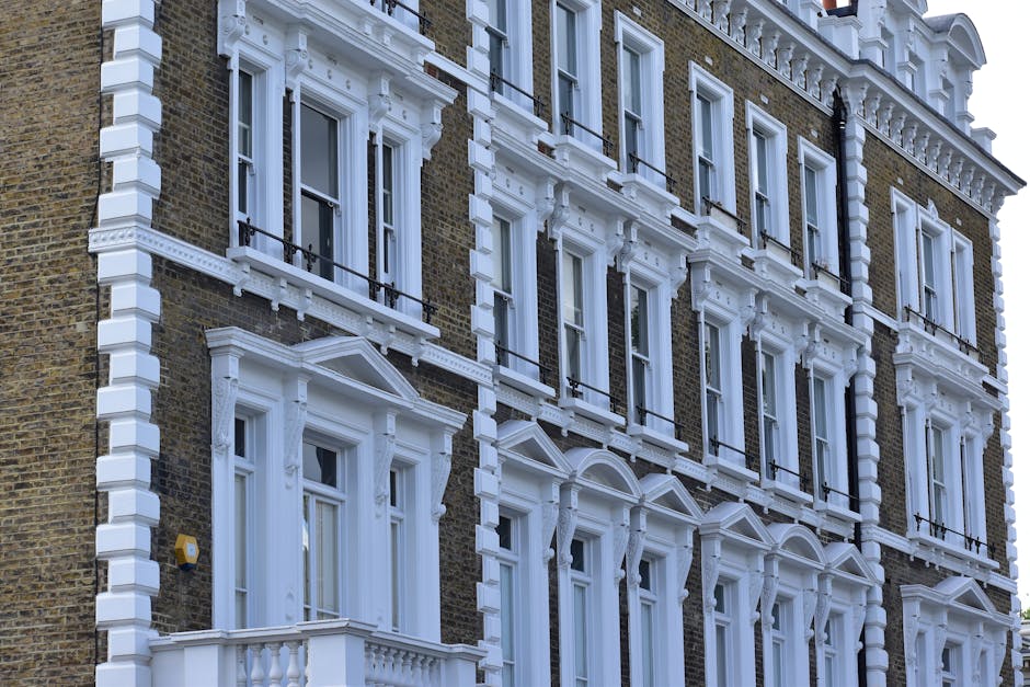 A multi-storey Victorian-style building with a dark brick façade and ornate white architectural details around the windows and along the cornice. The building features several large sash windows with black railings, and decorative white moldings above each window. The ground floor has a prominent white bay window with intricate trim and a small balustrade, while the upper floors display evenly spaced windows with similar white decorative framing. The exterior shows a clear contrast between the dark brickwork and the white embellishments, typical of traditional London townhouse architecture. This setting provides a typical urban environment where house removals and furniture transport services, such as those offered by Removal Van Marylebone, often take place during home relocation processes involving loading or unloading near the building entrance, under natural daylight.