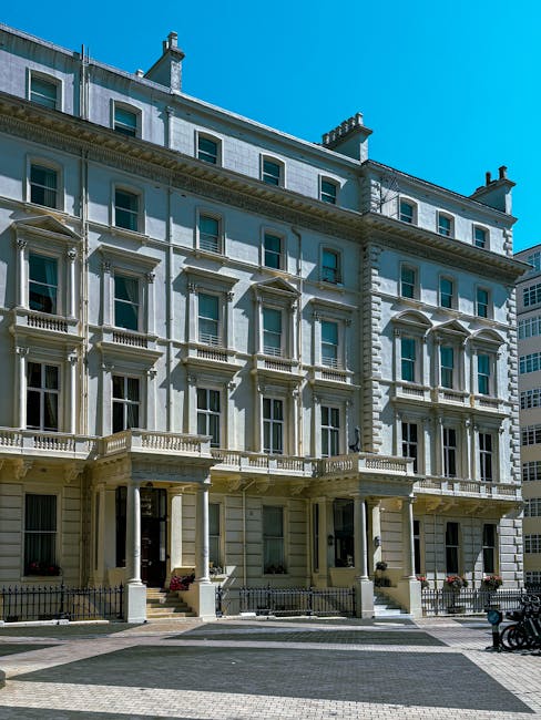 The image shows a multi-storey, white Victorian-style building with detailed architectural features and large windows, situated on a city street under a clear blue sky. In front of the building, there is a paved sidewalk with a low metal fence, some potted plants at the entrance, and a few bicycles parked nearby. The building's ground floor appears to have residential or commercial units. Although no moving activity is visible, the setting reflects the typical environment for house removals and furniture transport services offered by Removal Van Marylebone, including packing, loading, and logistical planning for home relocation projects in the Marylebone area.
