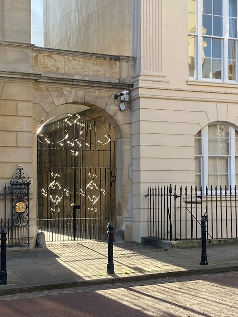 An ornate black metal gate with decorative patterns, set within a stone archway on the facade of a beige Georgian-style building. The building features large sash windows with white frames, detailed stonework, and a black wrought-iron fence running along the sidewalk. Shadows cast by the bollards and sunlight illuminate the pavement area in front of the entrance. The scene appears to be part of a property undergoing house removals, with potential access for furniture transport as part of a home relocation process. This setting reflects the typical environment for professional removals, as provided by Removal Van Marylebone, including parking considerations like Westminster Council permits.
