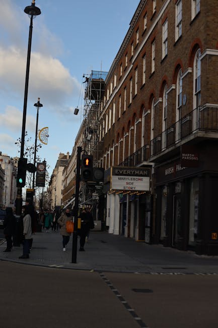 A street scene in Marylebone, Westminster, showing pedestrians walking along the pavement beneath a brick building with multiple floors and balconies. Some shop signs are visible, including a temporary banner for Removal Van Marylebone, indicating a nearby home relocation service. In the foreground, a traffic light displays a red signal for pedestrians, and a street lamp is seen rising beside the sidewalk. The building's upper floors are under scaffolding and construction netting, suggesting ongoing building maintenance or renovation. The photograph captures a typical urban environment with a mixture of retail storefronts and residential lodges, suitable for facilitating household moves or furniture transport for local removals companies.